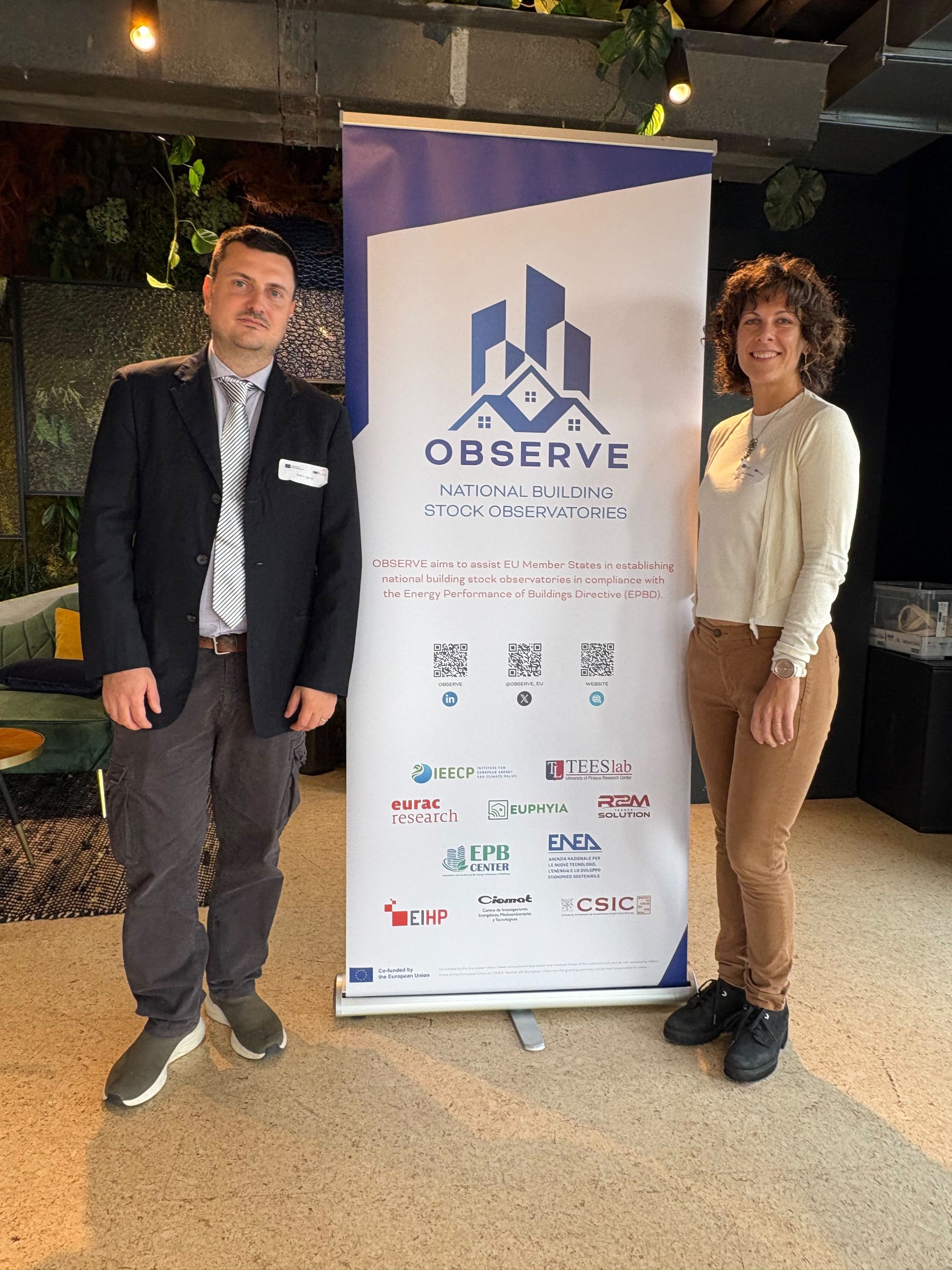Fotografia scattata durante un evento del progetto OBSERVE. Fabio Cignini e Francesca Pagliaro di ENEA sono in piedi ai lati di un banner verticale che riporta il logo del progetto e le informazioni sull'iniziativa europea "National Building Stock Observatories". Sul roll-up sono visibili il nome del progetto, alcuni codici QR e i loghi dei partner, tra cui ENEA. L'immagine documenta la partecipazione all'evento dedicato al miglioramento della raccolta, della qualità e dell'interoperabilità dei dati sul patrimonio edilizio europeo