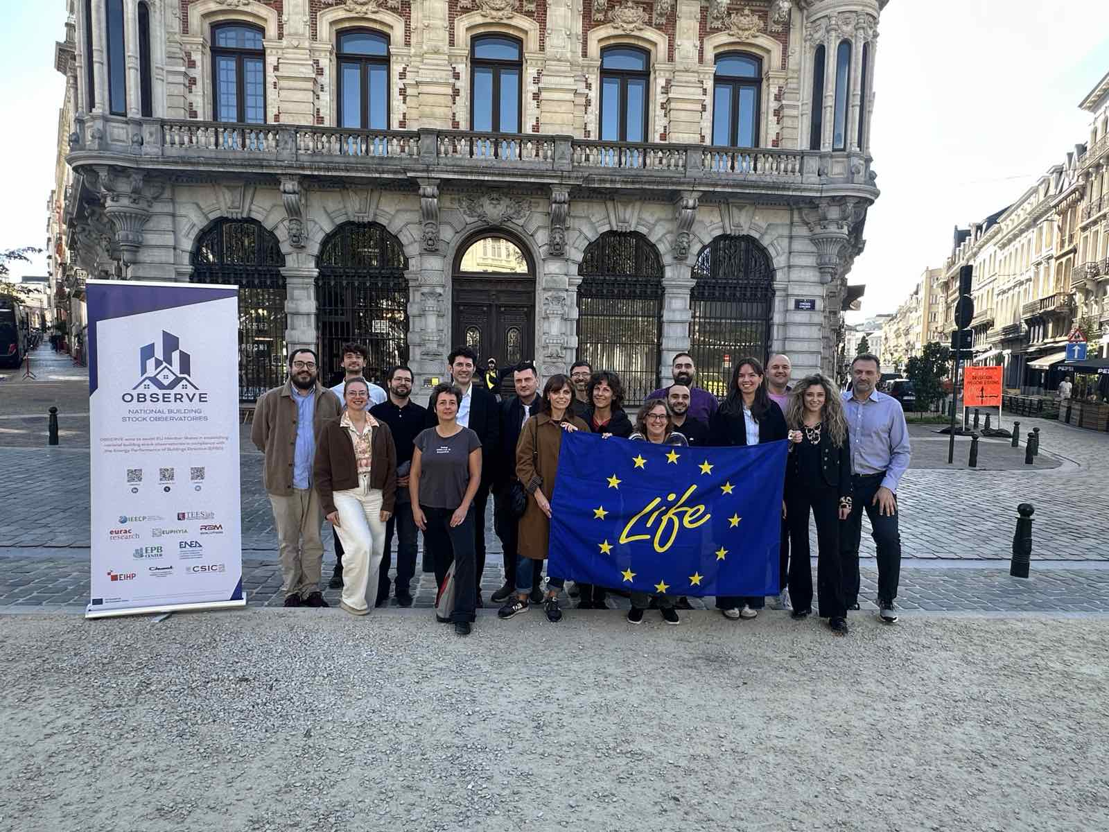 Fotografia realizzata a Bruxelles durante un incontro del progetto OBSERVE (National Building Stock Observatories). Un gruppo di partecipanti provenienti dalle organizzazioni partner è riunito all'aperto davanti a un edificio storico. Alla sinistra del gruppo è collocato un roll-up con il logo e le informazioni del progetto OBSERVE, mentre al centro è esposta una bandiera del programma europeo LIFE. L'immagine documenta la collaborazione internazionale nell'ambito delle attività finalizzate a migliorare la raccolta, la qualità e l'interoperabilità dei dati sul patrimonio edilizio europeo