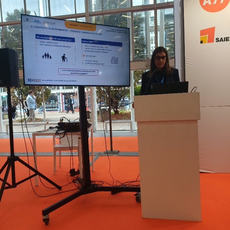 All'interno di uno stand del SAIE di Bologna, Elisabetta Parisi (ENEA) interviene da un podio davanti a un monitor su supporto mobile. Sullo schermo è visibile una slide del progetto REHOUSE dedicata al contesto della povertà energetica negli edifici di edilizia residenziale pubblica (ERP)
